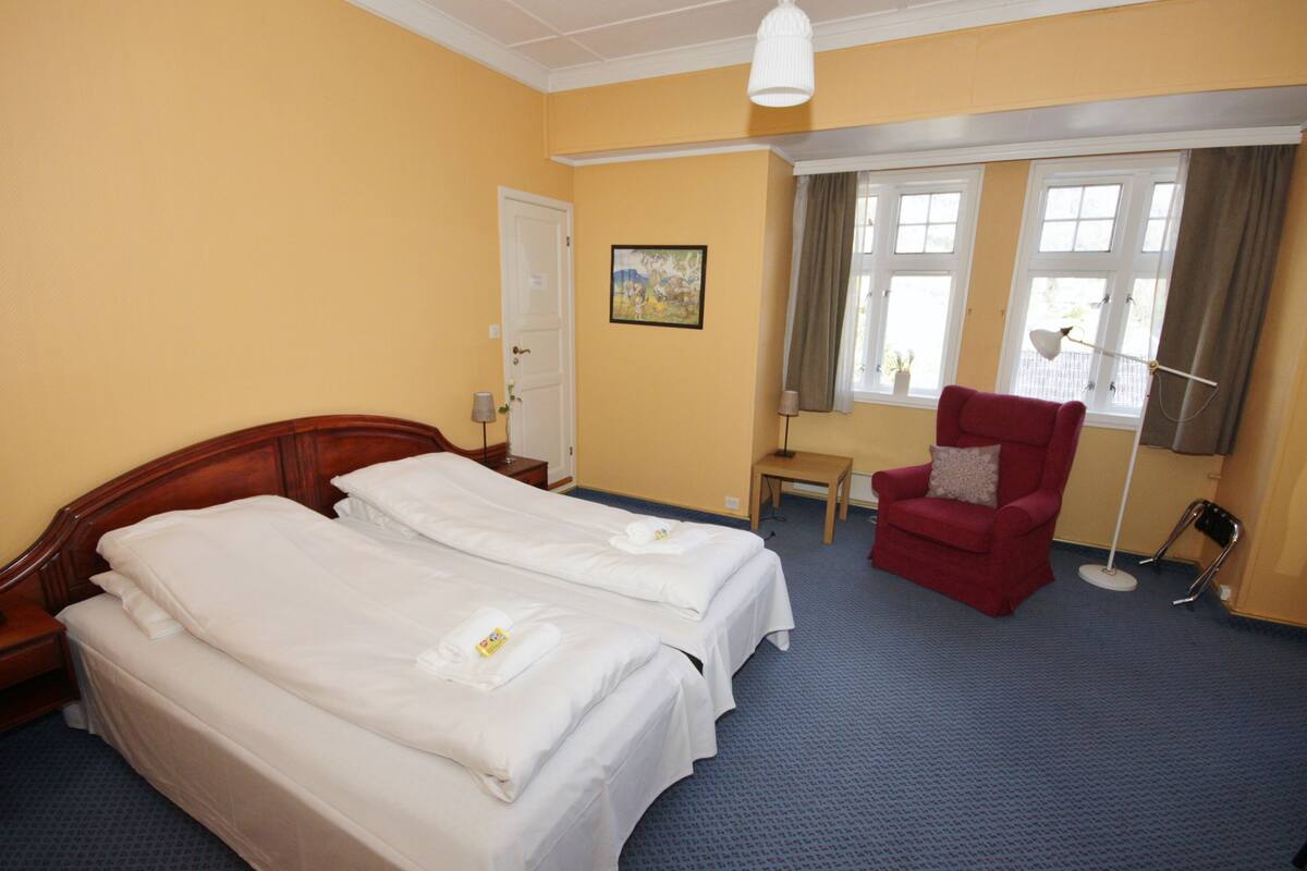 A hotel room with two beds, a red armchair, a small table, and a window with curtains.