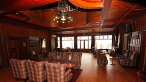 A spacious room with wooden floors and walls, featuring multiple seating areas with plaid-patterned sofas and armchairs, a chandelier, and large windows providing natural light. - Sauda Fjordhotell (Sauda)