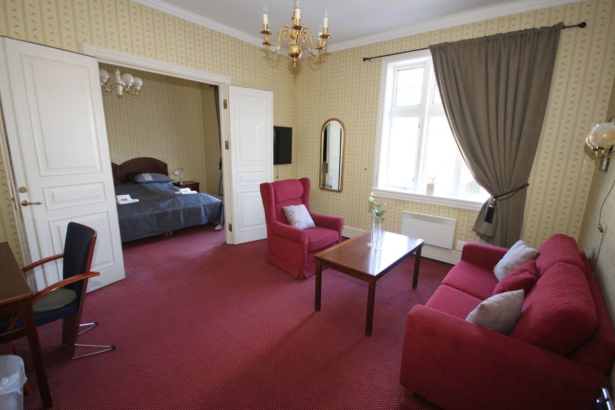 A hotel room with a bed, a red armchair, a sofa, a wooden coffee table, and a large mirror.