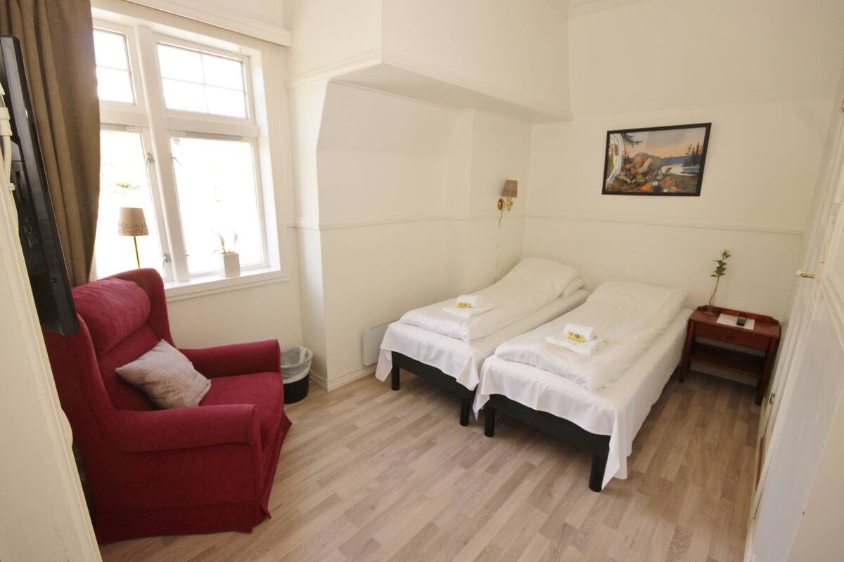 A room with two beds, a red armchair, a window with curtains, a painting on the wall, and a small table with a plant.