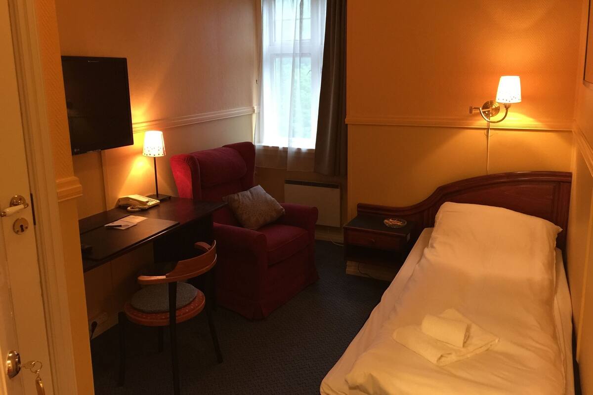 A hotel room with a bed, a desk with a telephone, a chair, a small table with a lamp, and a television mounted on the wall.