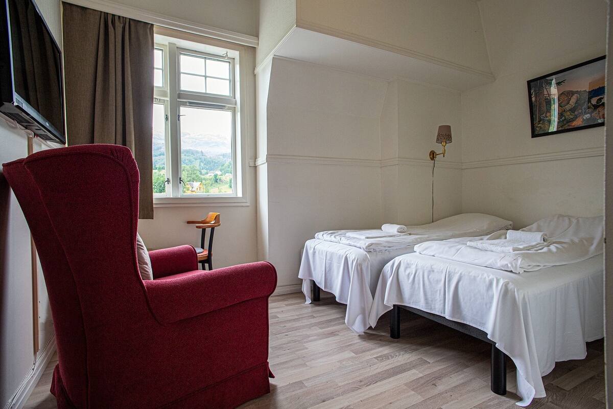 A hotel room with two beds, a red armchair, a window with a view, and a painting on the wall.