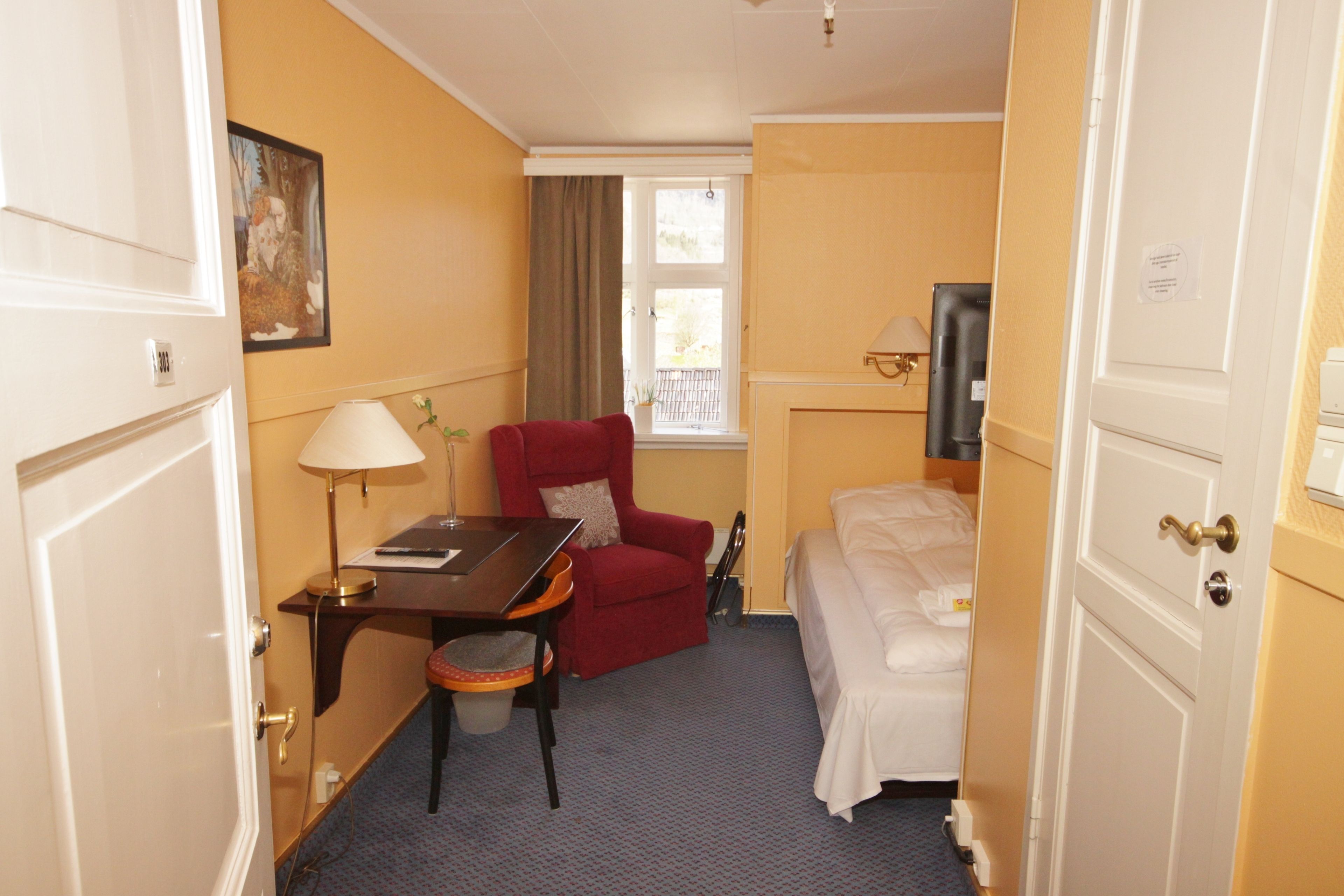 A hotel room with a bed, a desk with a chair, a red armchair, a television, and a window with curtains.