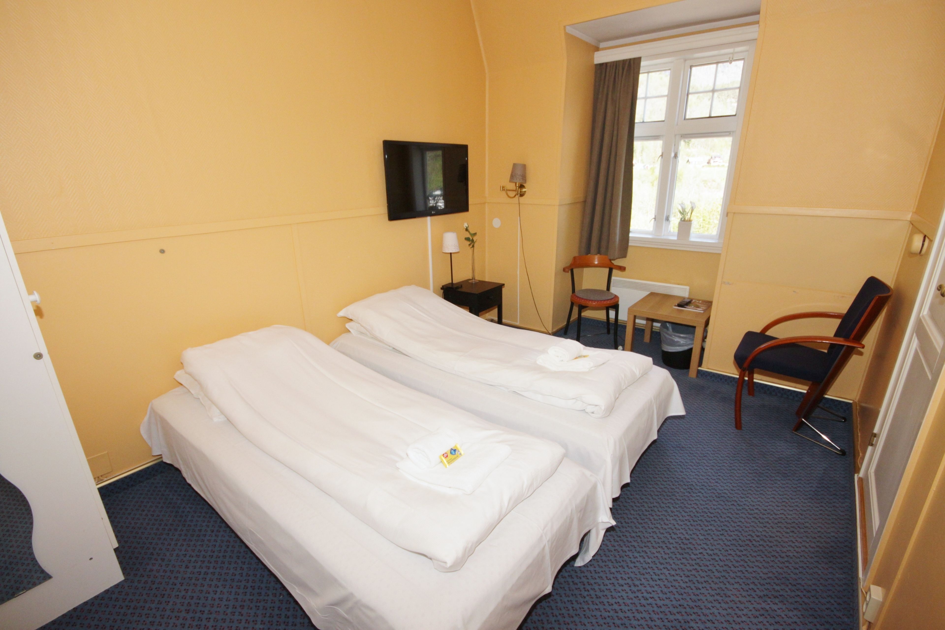 Une chambre d’hôtel avec deux lits simples, un bureau avec une chaise, un téléviseur fixé au mur et une fenêtre avec des rideaux.