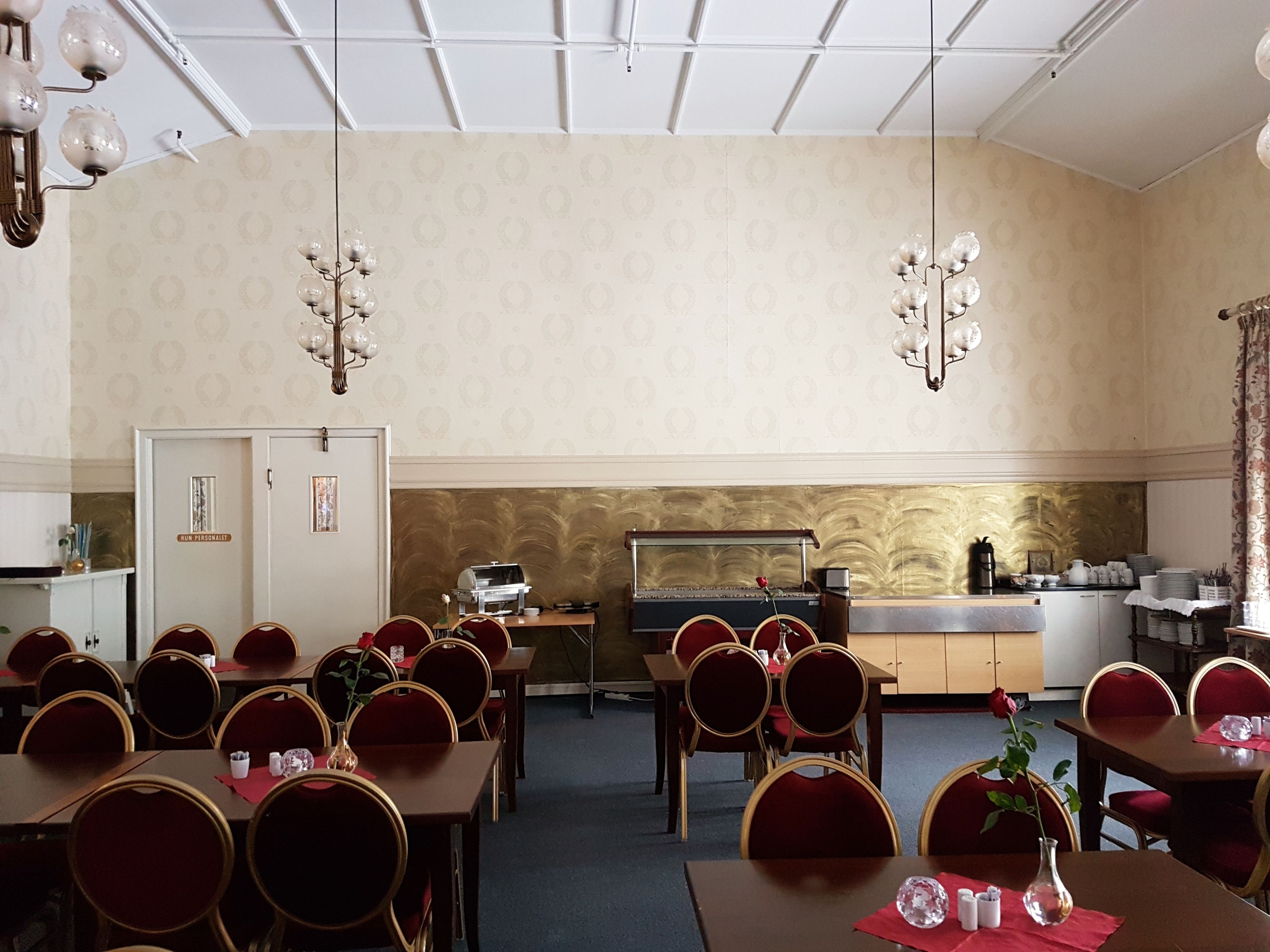 Un espace repas avec plusieurs tables et chaises, une cuisine à l’arrière-plan et un plafond orné d’un motif géométrique.