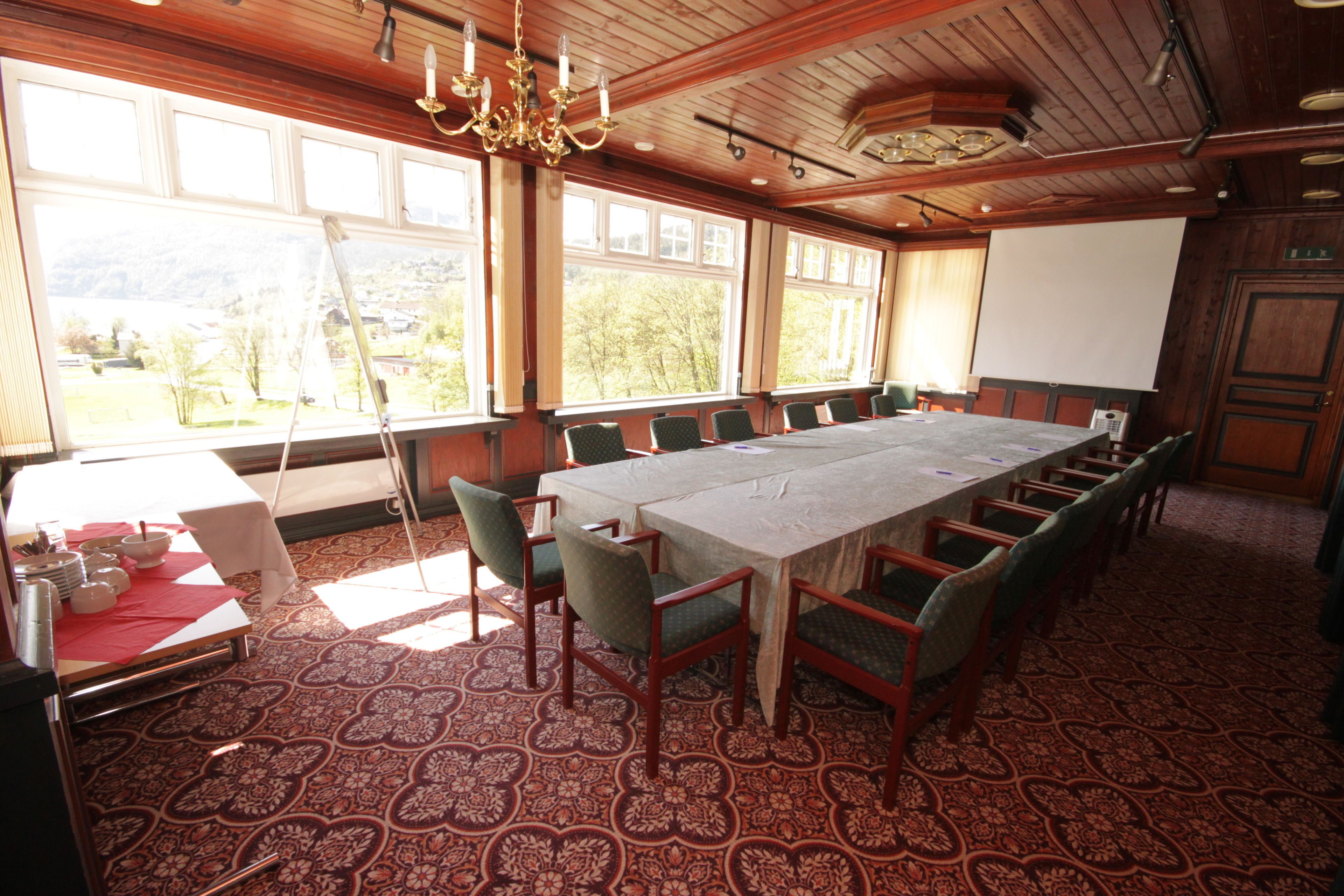 A conference room with a long table, chairs, a projector screen, and large windows with a view of greenery.