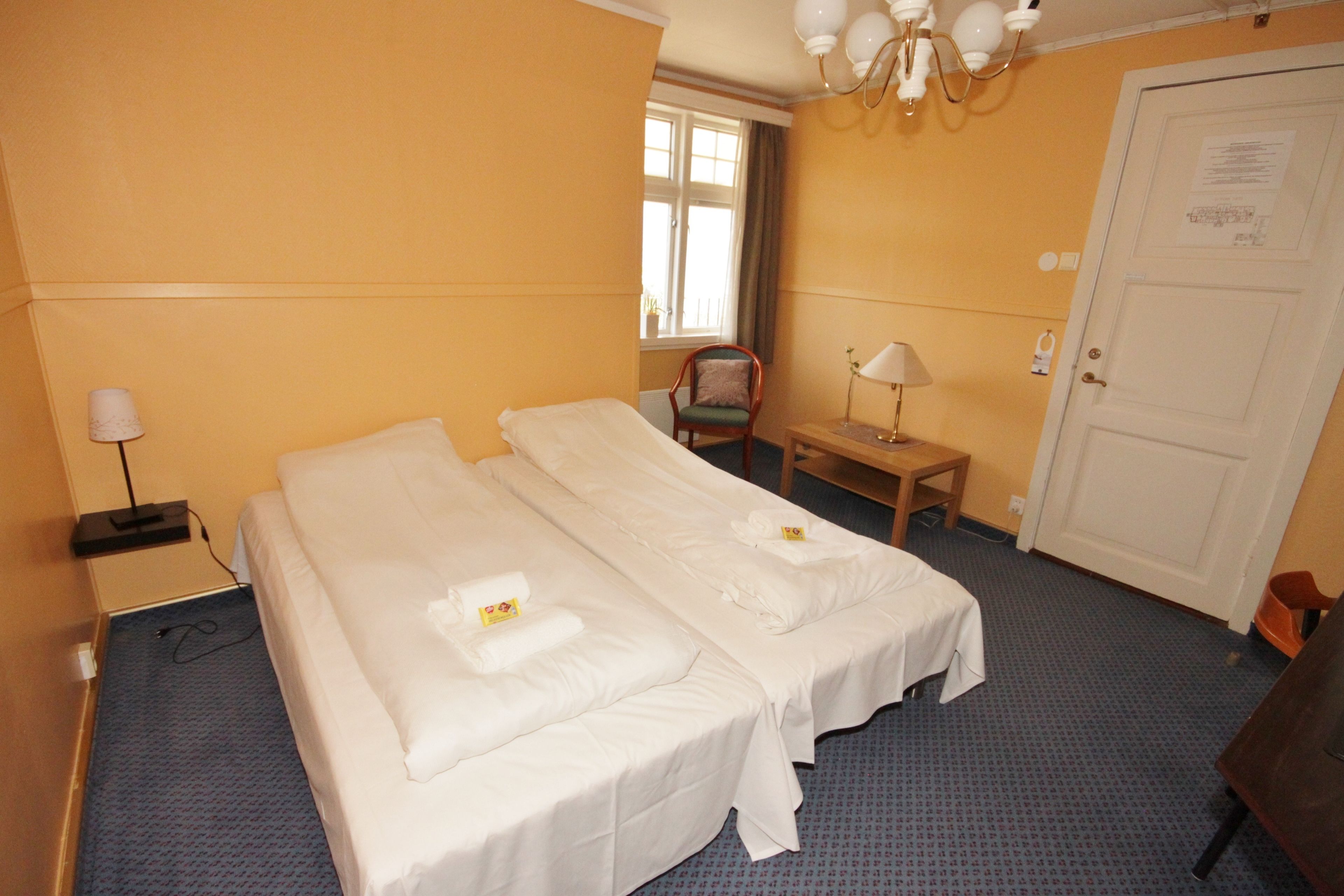 A hotel room with two beds, a chair, a desk, a lamp, and a door.
