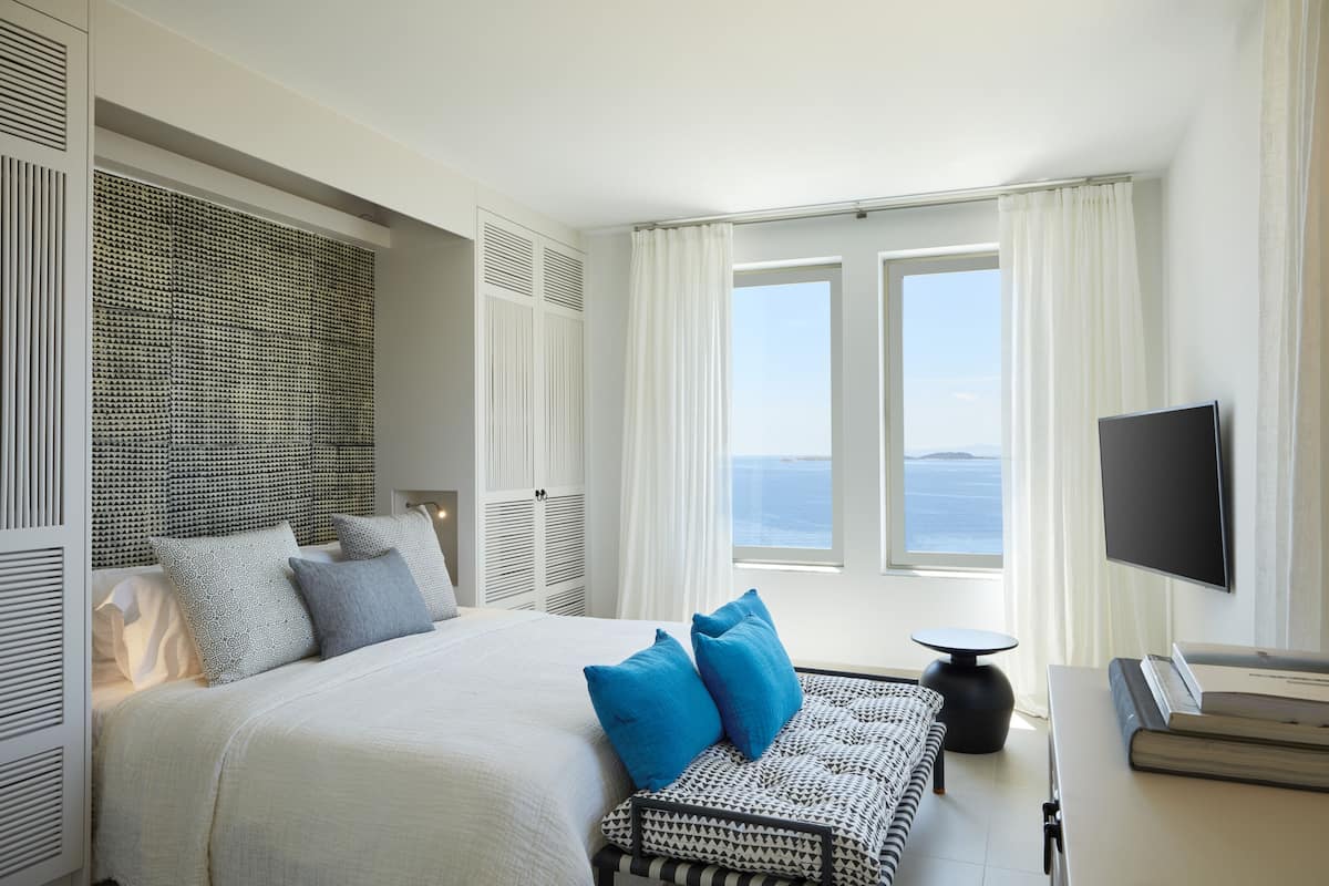 Ein Hotelzimmer mit einem großen Bett, einem Flachbildfernseher, Blick auf das Meer und einem Fenster mit weißen Vorhängen.