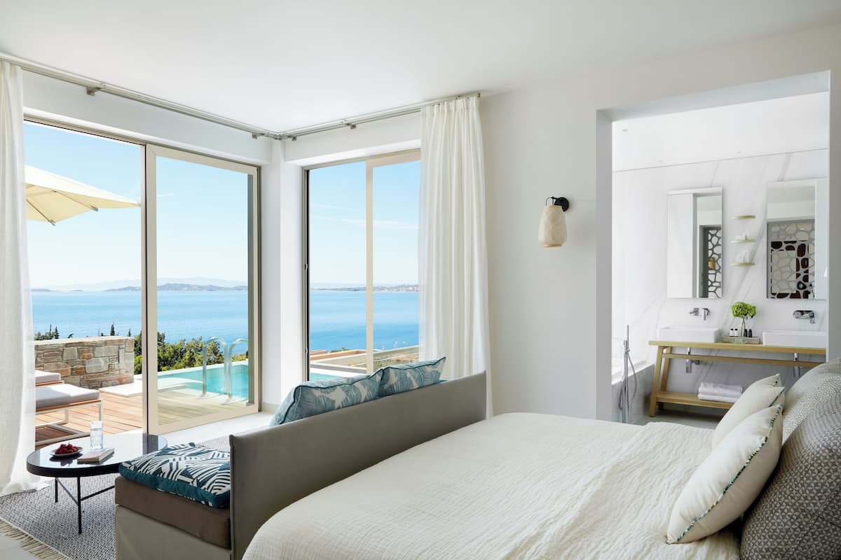 Ein Hotelzimmer mit Bett, kleinem Tisch, Sofa, Badezimmer mit Spiegel und Meerblick.