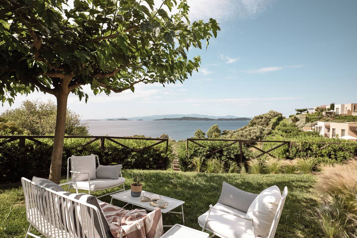 Eine Terrasse mit zwei weißen Stühlen und einem Tisch, mit Blick auf ein Gewässer und Hügel.