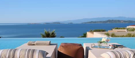 A poolside lounge with two cushioned chairs, a glass of wine, and a book on a table, overlooking a clear blue sea and distant land.