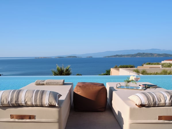 A poolside lounge with two cushioned chairs, a glass of wine, and a book on a table, overlooking a clear blue sea and distant land.