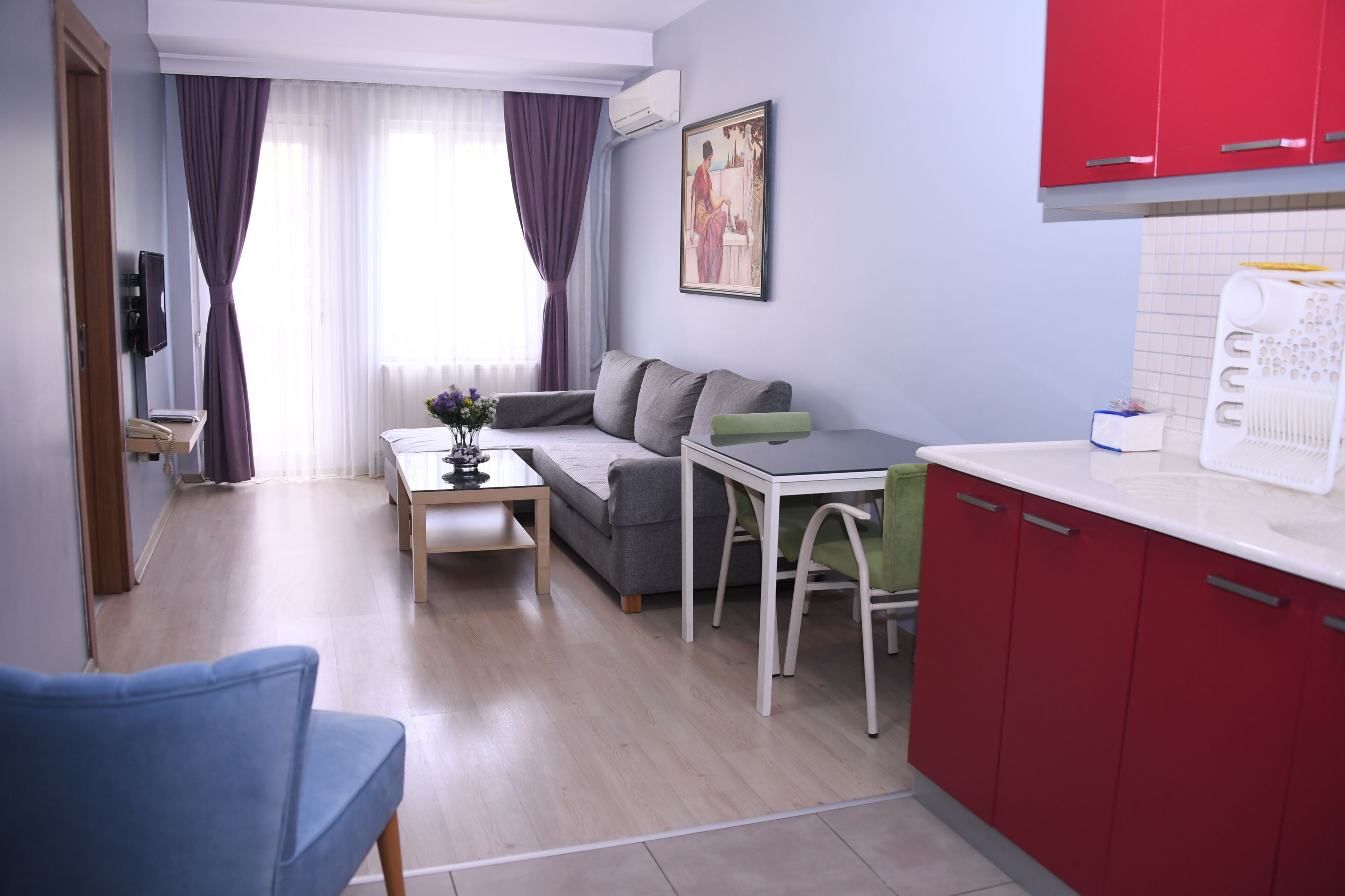 Espaço compacto com sofá, mesa de jantar, área de cozinha com armários vermelhos e janela com cortinas.