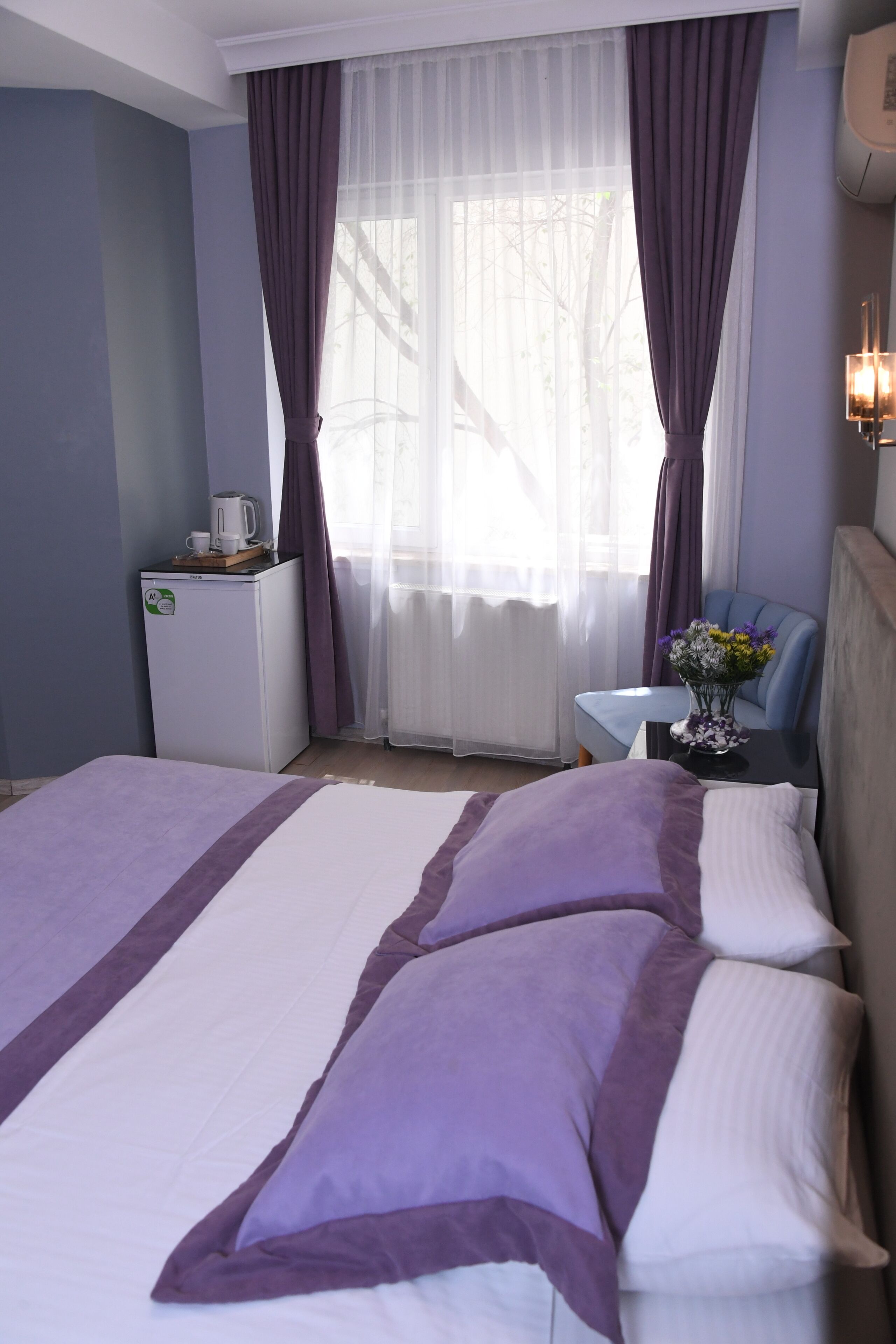 Una habitación de hotel con cama, una mesita con un jarrón de flores, una cafetera y una ventana con cortinas.