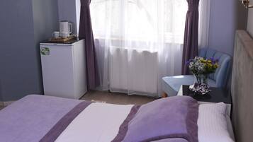Una habitación de hotel con cama, una mesita con un jarrón de flores, una cafetera y una ventana con cortinas.