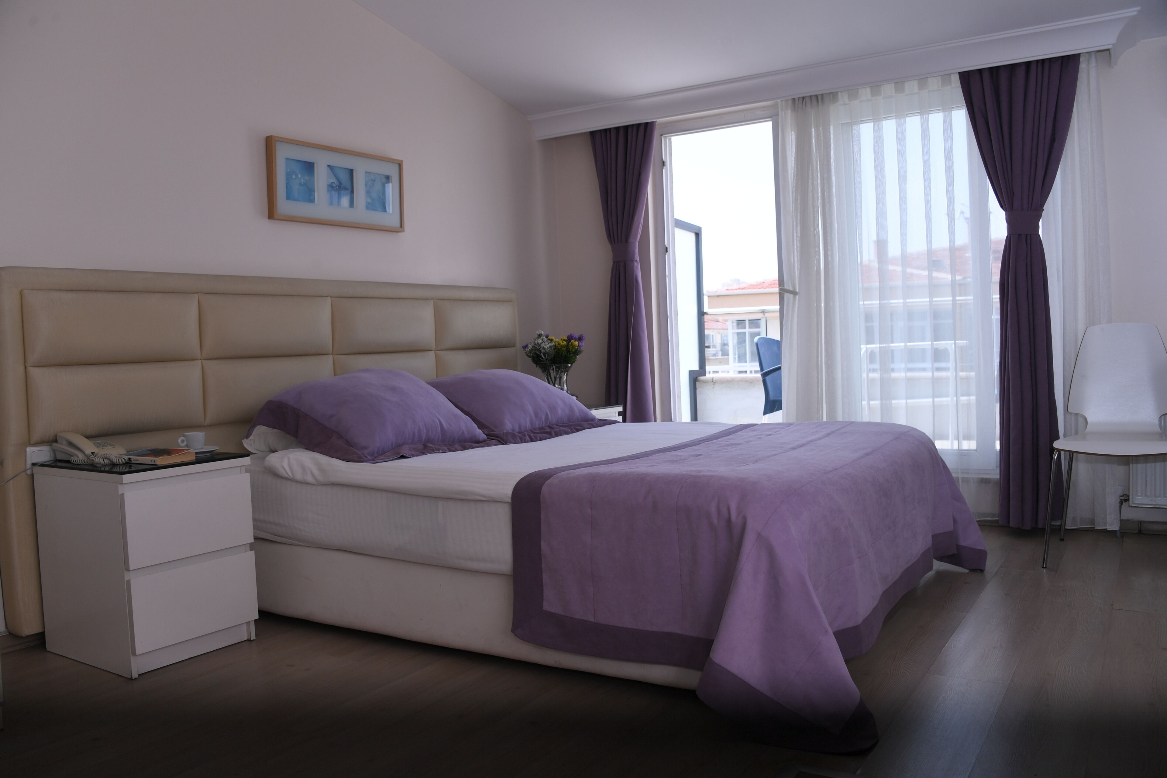 Een hotelkamer met een bed, nachtkastje, stoel en een raam met gordijnen.