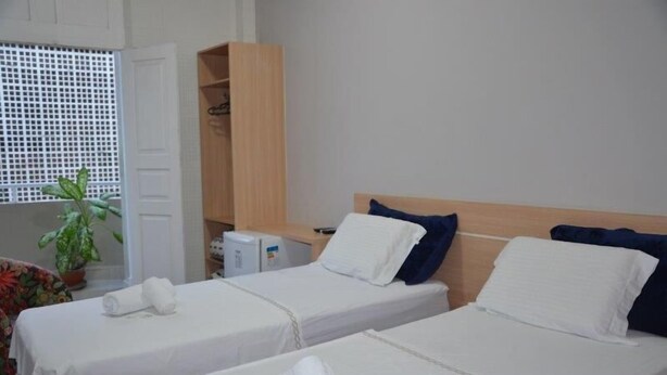 A hotel room with two single beds, a wooden wardrobe, a small table with a plant, and a window with a white lattice screen. - Sumare Hotel (Florianópolis)