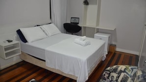 A neatly made bed with white linens, a bedside table with a lamp, a chair, and a small shelf. - Sumare Hotel (Florianópolis)