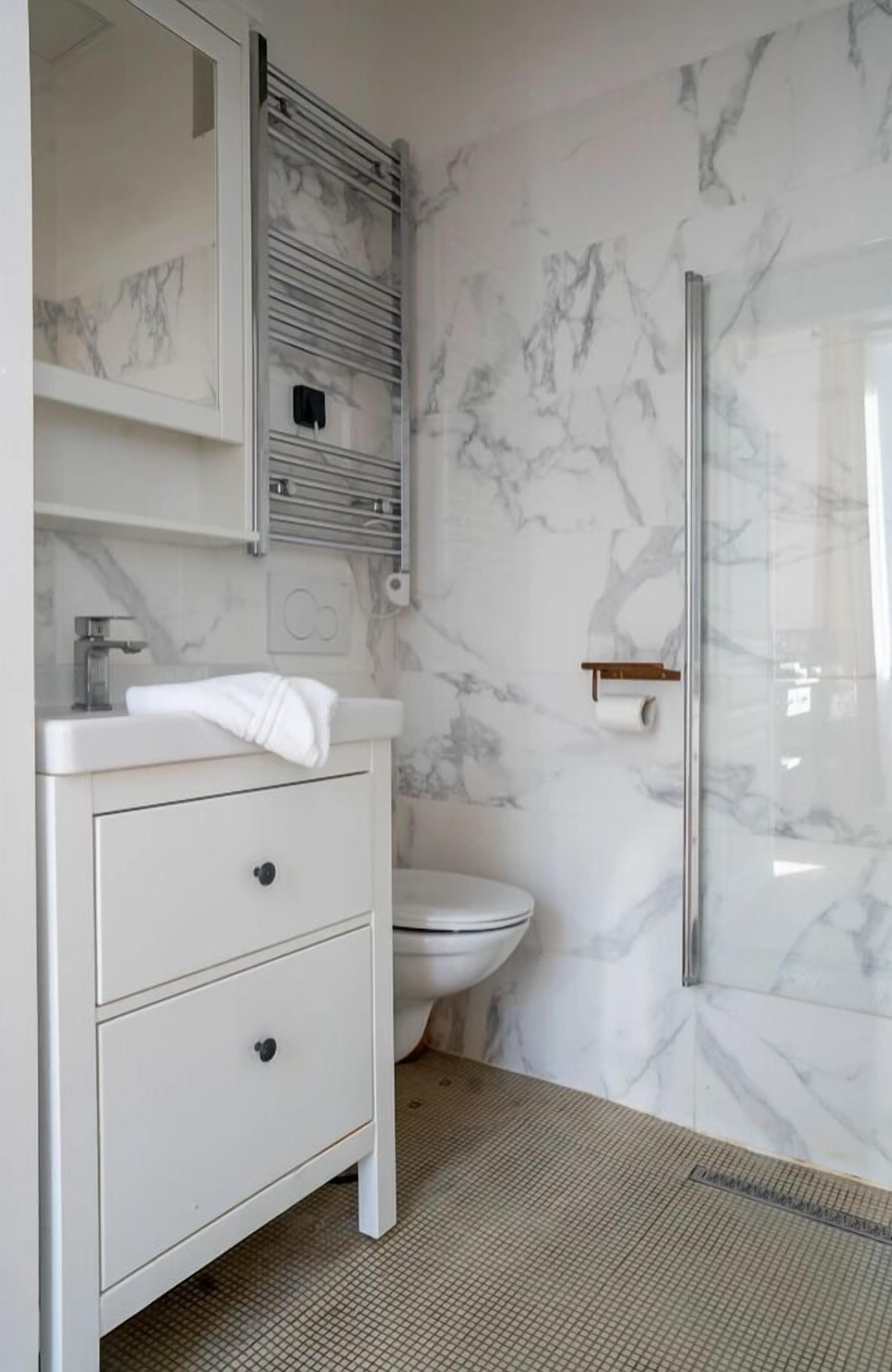 A modern bathroom with a white vanity, marble walls, a glass shower enclosure, and a toilet.