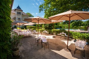 An outdoor patio with tables and chairs, umbrellas, and a pool in the background.