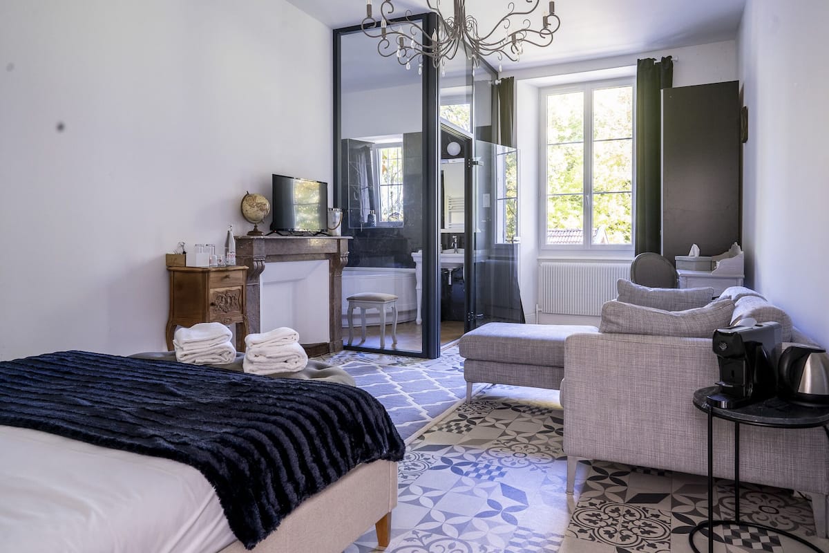 A bedroom with a bed, a desk, a chair, a sofa, and a bathroom visible through a glass door.