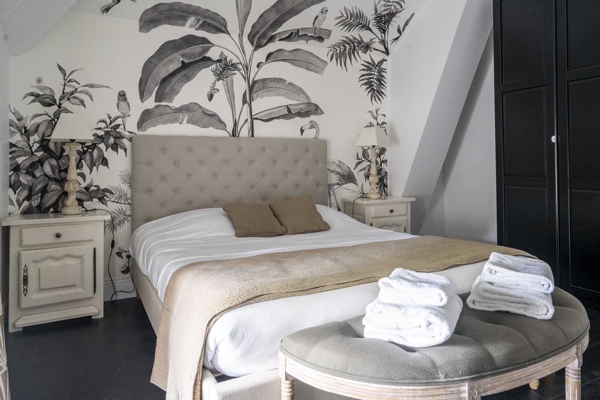 Ein Schlafzimmer mit einem großen Bett, einem Kopfteil, zwei Nachttischen, einer Bank mit Handtüchern und einem Wandbild.