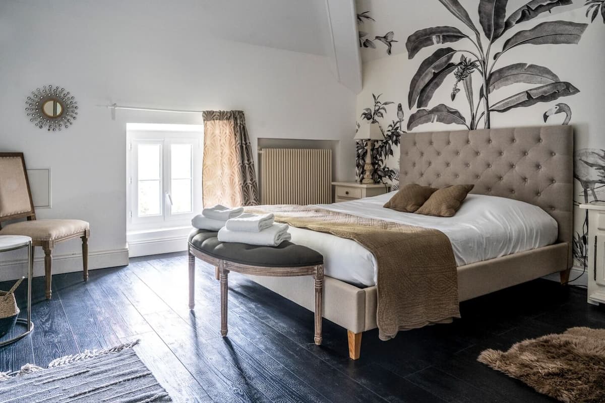 A bedroom with a large bed, a round bench, a chair, and a wall with a tropical mural.