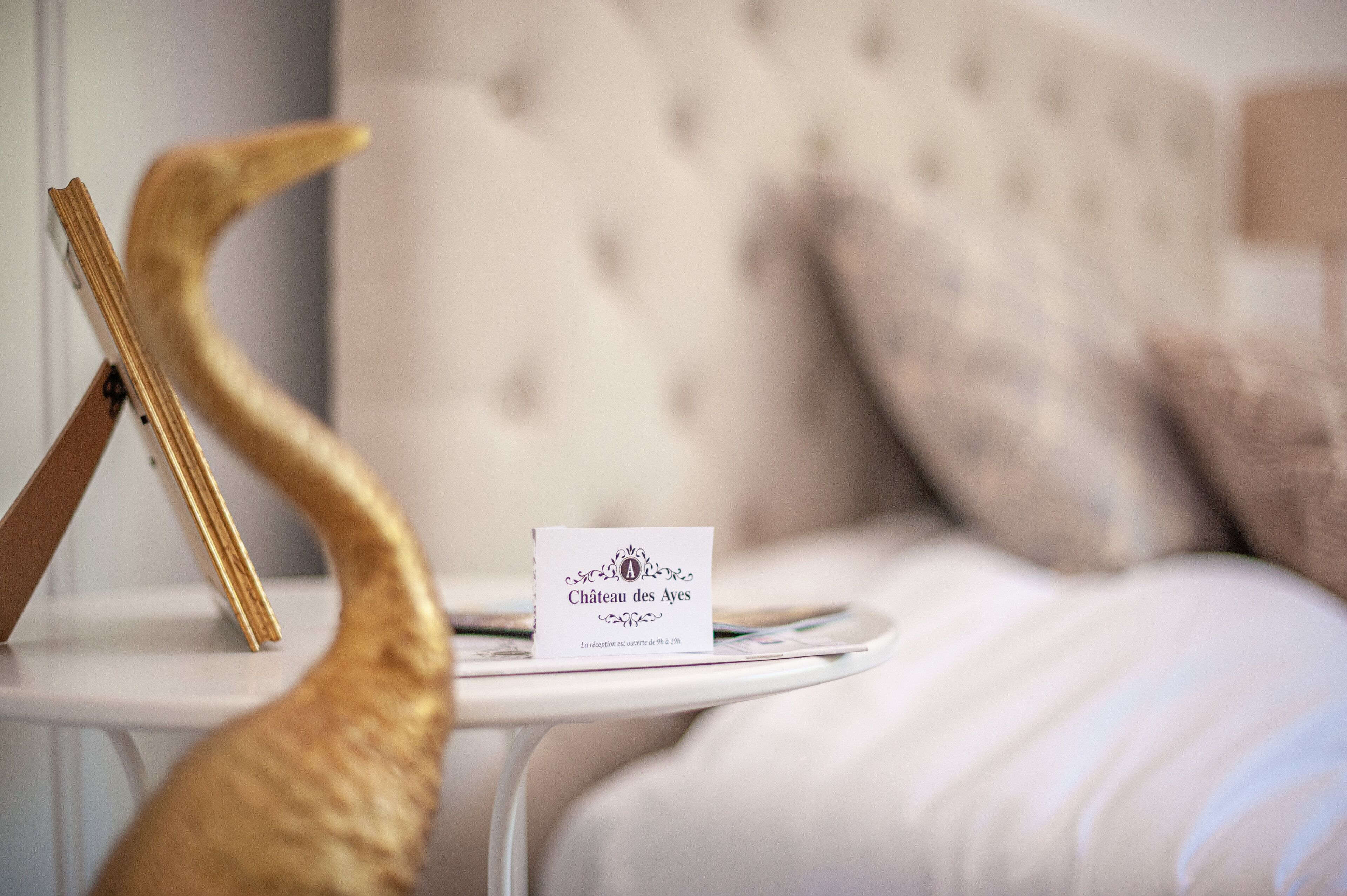 A bed with a tufted headboard, a white bedside table with a sign reading "Château des Ayes," and a golden decorative object.
