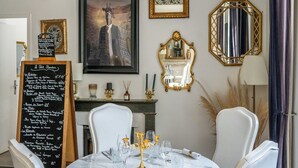 A dining area with a round table set for four, white chairs, a gold chandelier, and a blackboard menu. - Château Des Ayes (Saint-Etienne-de-Saint-Geoirs)