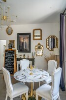 A dining area with a round table set for four, white chairs, a gold chandelier, and a blackboard menu.