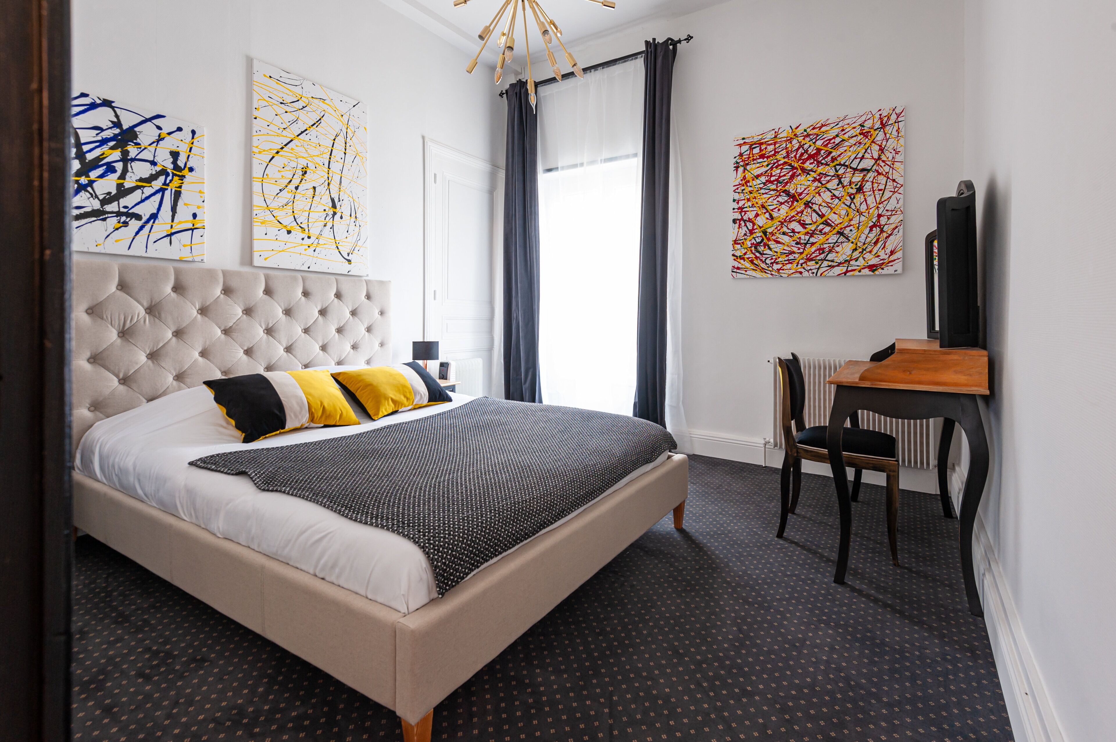 Un dormitorio con cabecera capitonada, cama con ropa de cama blanca y negra, un escritorio con silla y arte abstracto en las paredes.