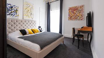 Un dormitorio con cabecera capitonada, cama con ropa de cama blanca y negra, un escritorio con silla y arte abstracto en las paredes.