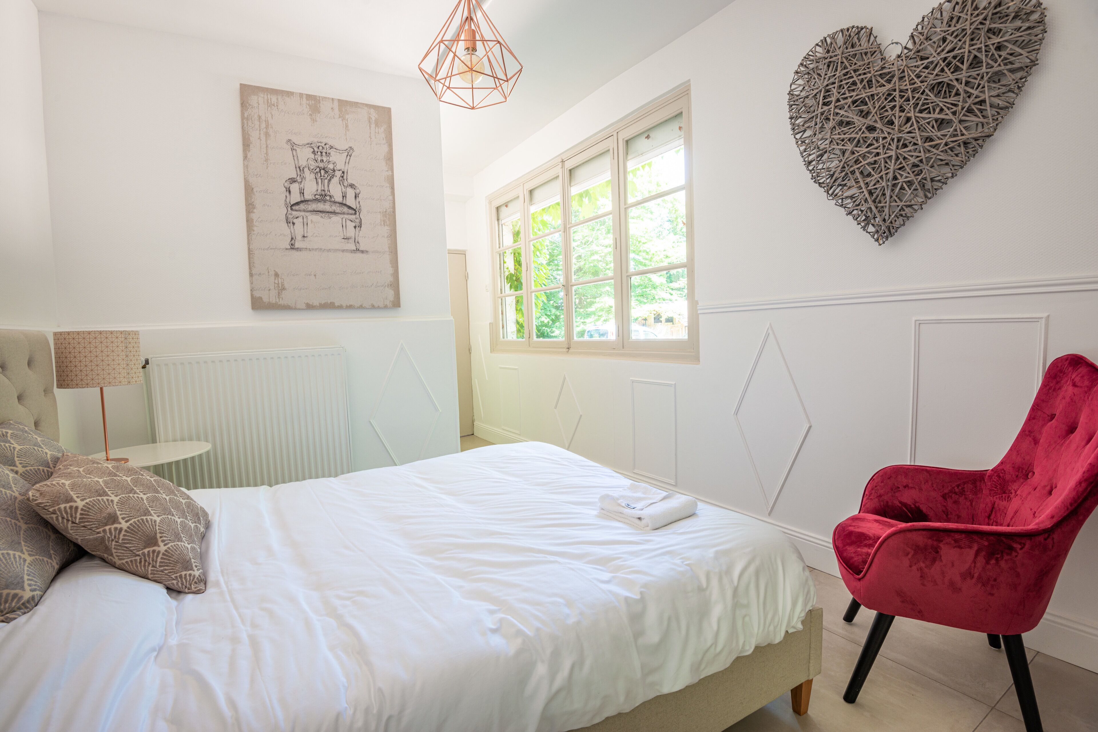A bedroom with a bed, a red chair, a window, and a wall art piece.