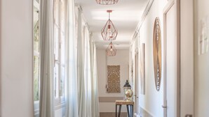 A hallway with a tiled floor, white walls, and a door on the right. There are curtains on the left side, a table with a lamp, and a hanging light fixture. - Château Des Ayes (Saint-Etienne-de-Saint-Geoirs)