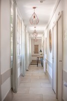 A hallway with a tiled floor, white walls, and a door on the right. There are curtains on the left side, a table with a lamp, and a hanging light fixture.