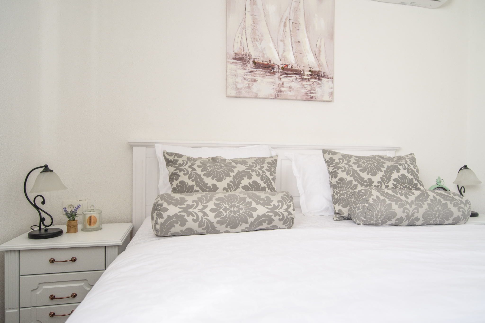 A neatly made bed with floral-patterned pillows, a bedside table with a lamp and a small plant, and a wall-mounted painting of sailboats.
