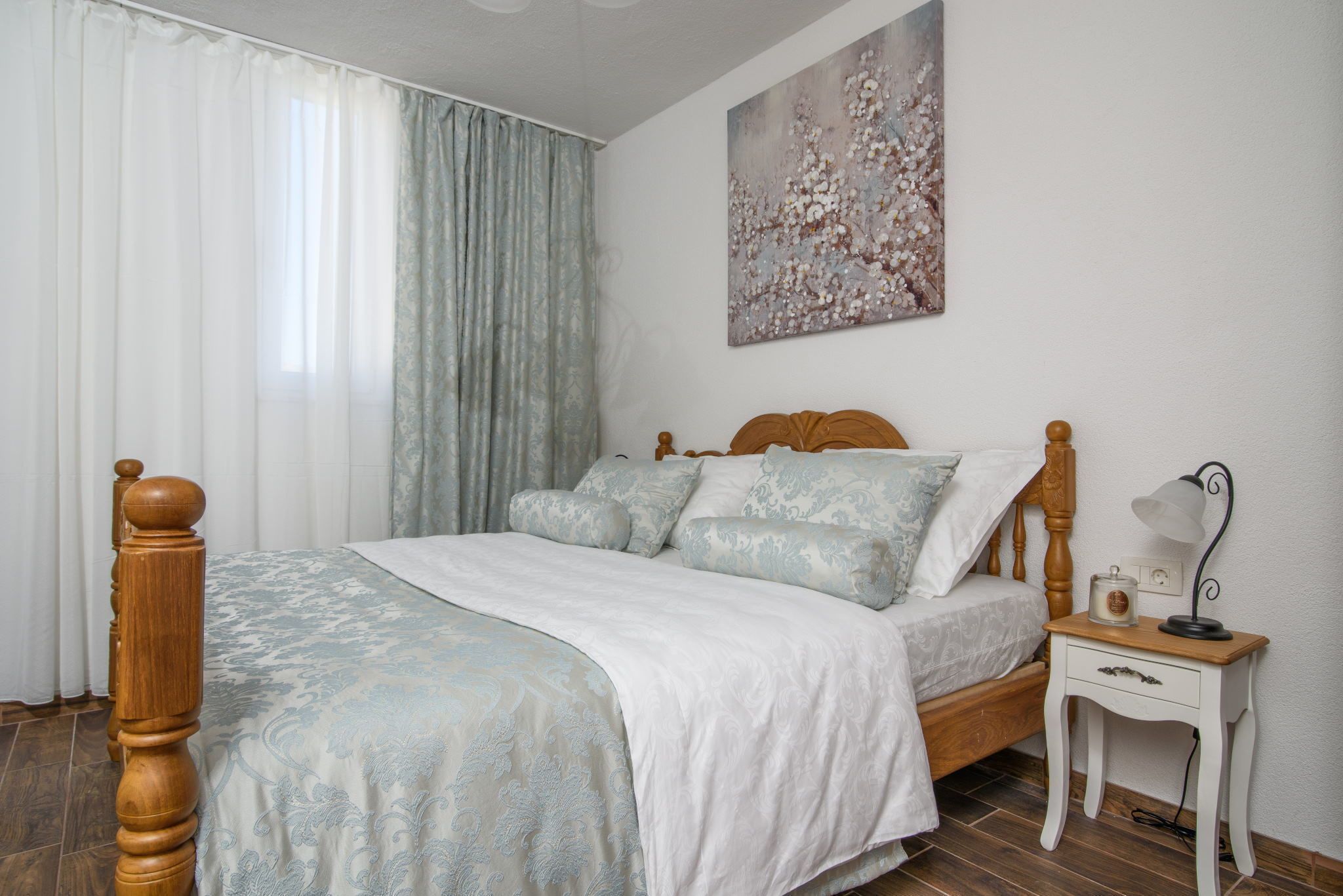 Un dormitorio con una cama de madera, ropa de cama blanca, una mesita de noche con una lámpara y un cuadro floral en la pared.