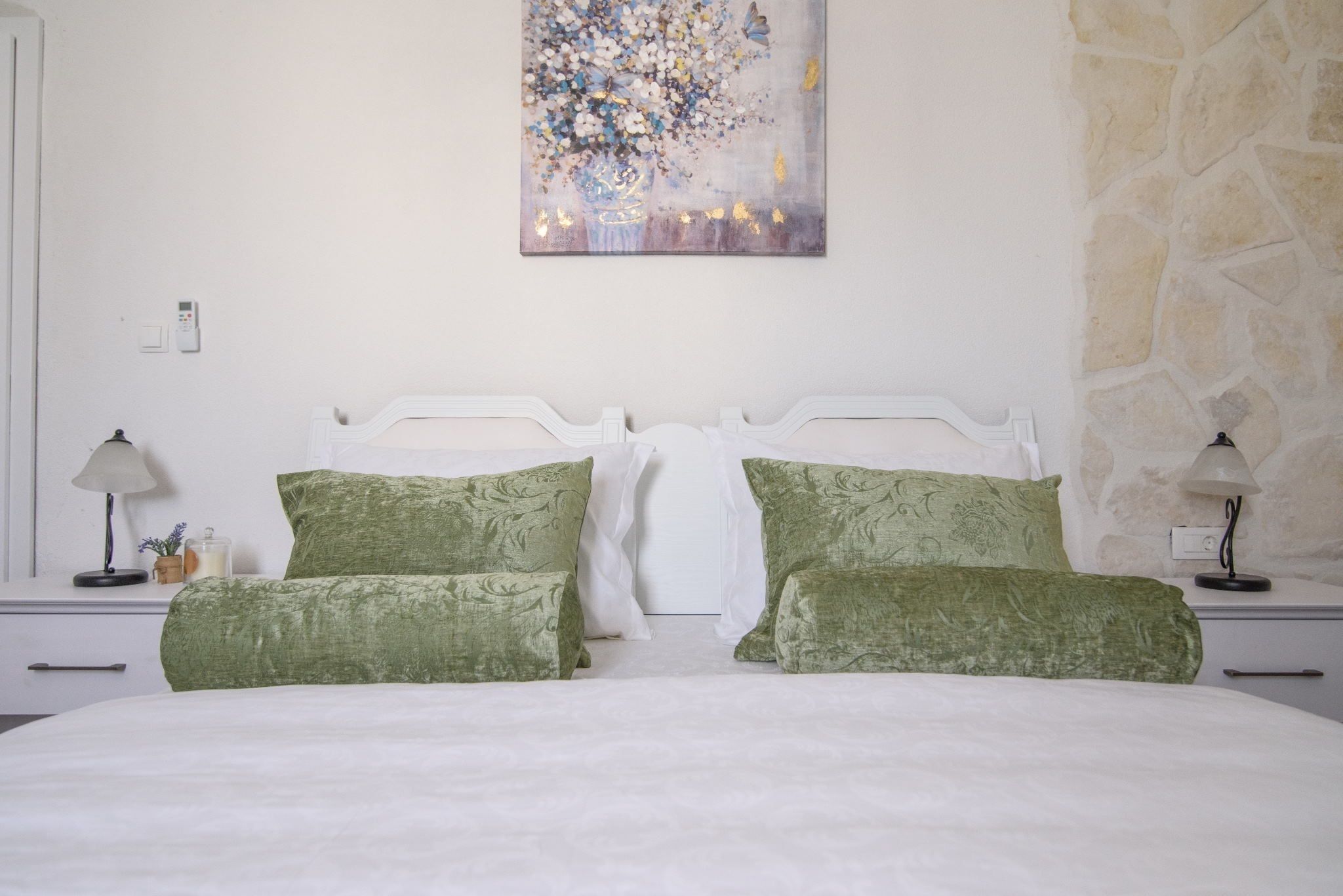 Une chambre avec un lit blanc, des oreillers verts, un tableau au mur et un mur en pierre.
