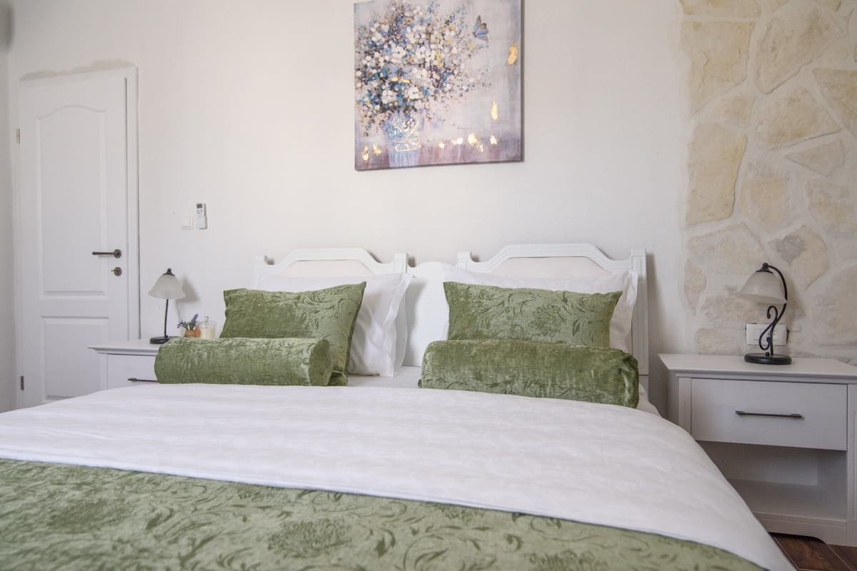 Un dormitorio con cama blanca, almohadas verdes, una mesita de noche con lámpara y un cuadro en la pared.