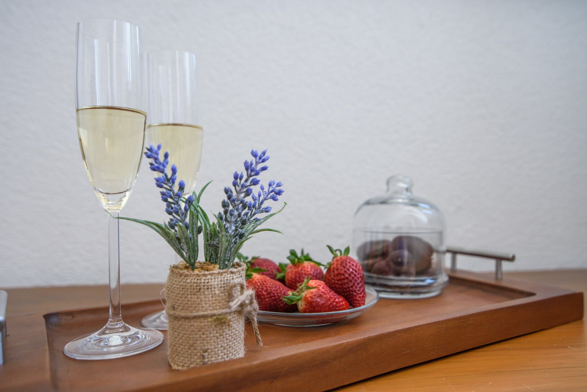 Two glasses of white wine, a basket of strawberries, and a glass jar with lavender on a wooden tray.