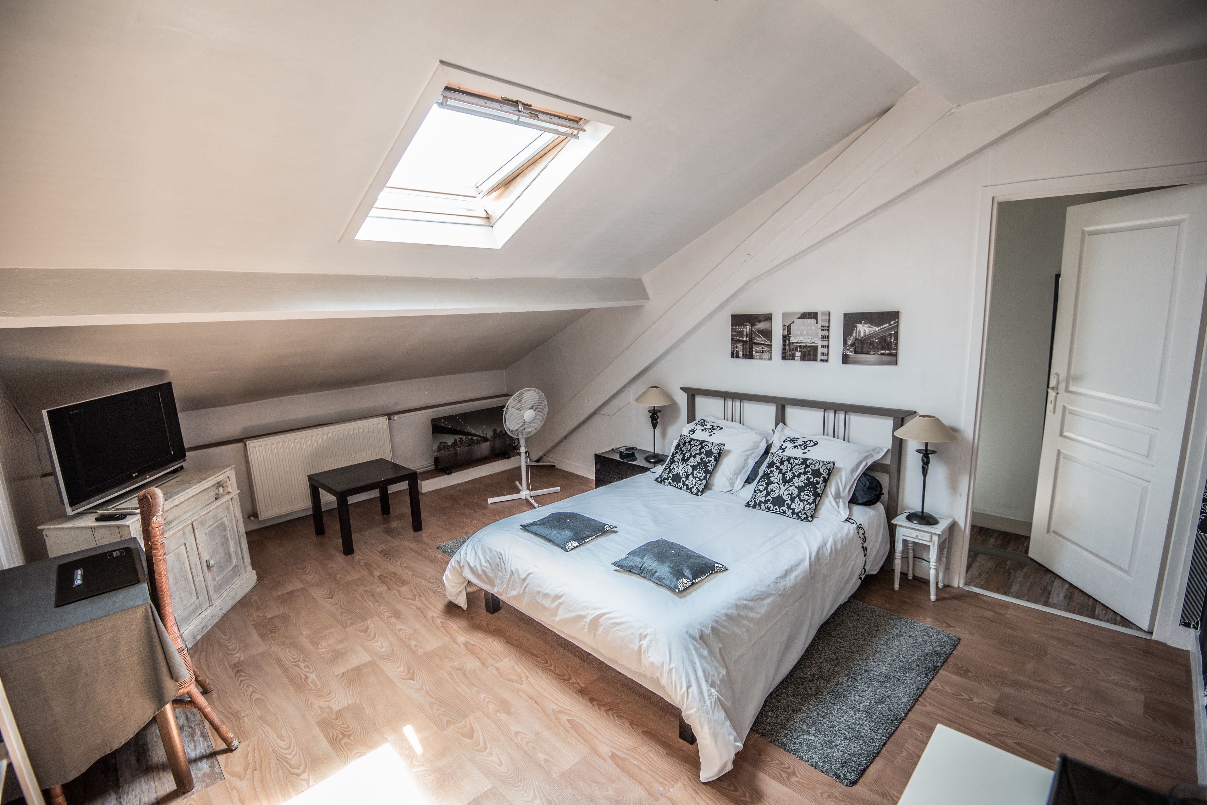 A bedroom with a skylight, a bed with pillows, a desk with a computer, a chair, and a small table with a lamp.