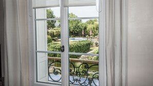 Uitzicht door een raam met een witte lijst en jaloezieën, op een tuin met een zwembad, een houten terras en een metalen balustrade.