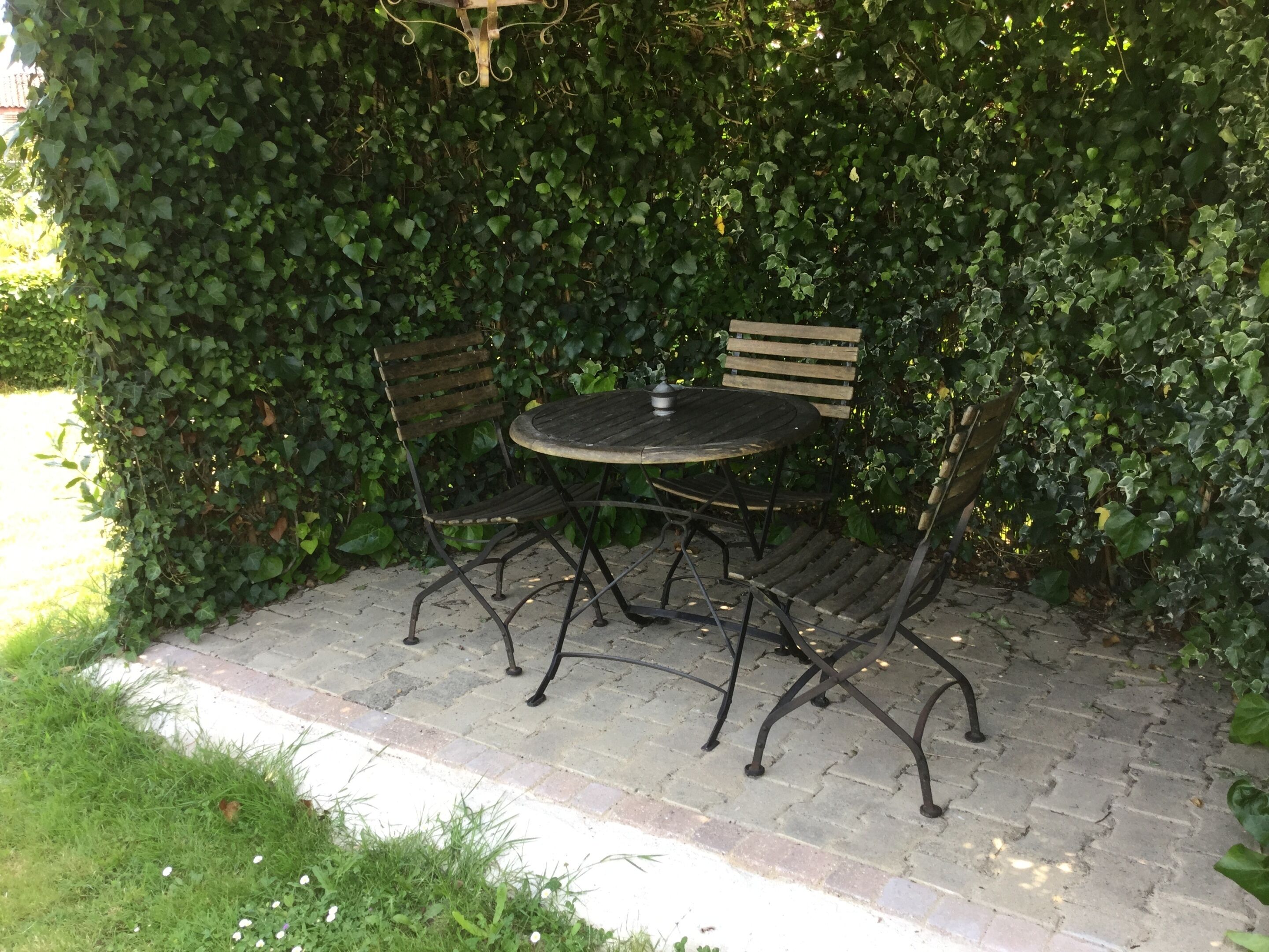 Uma área externa com espaço para sentar, incluindo uma mesa redonda e duas cadeiras, cercada por um muro verde denso.