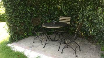 Uma área externa com espaço para sentar, incluindo uma mesa redonda e duas cadeiras, cercada por um muro verde denso.