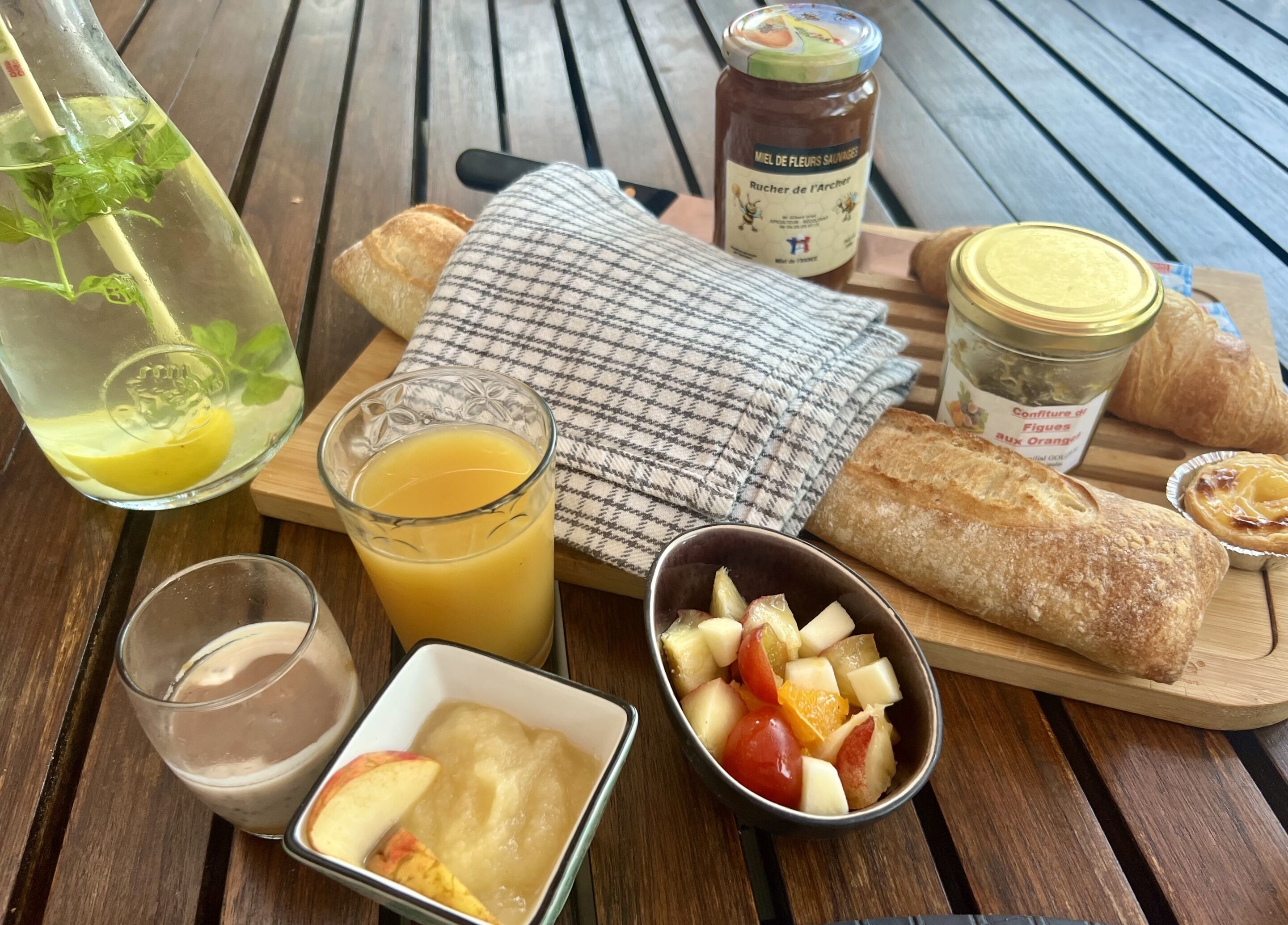 Un tavolo di legno con un bicchiere di succo d'arancia, una bottiglia di miele, un vasetto di marmellata, una baguette, una piccola ciotola di frutta e un bicchiere di latte.