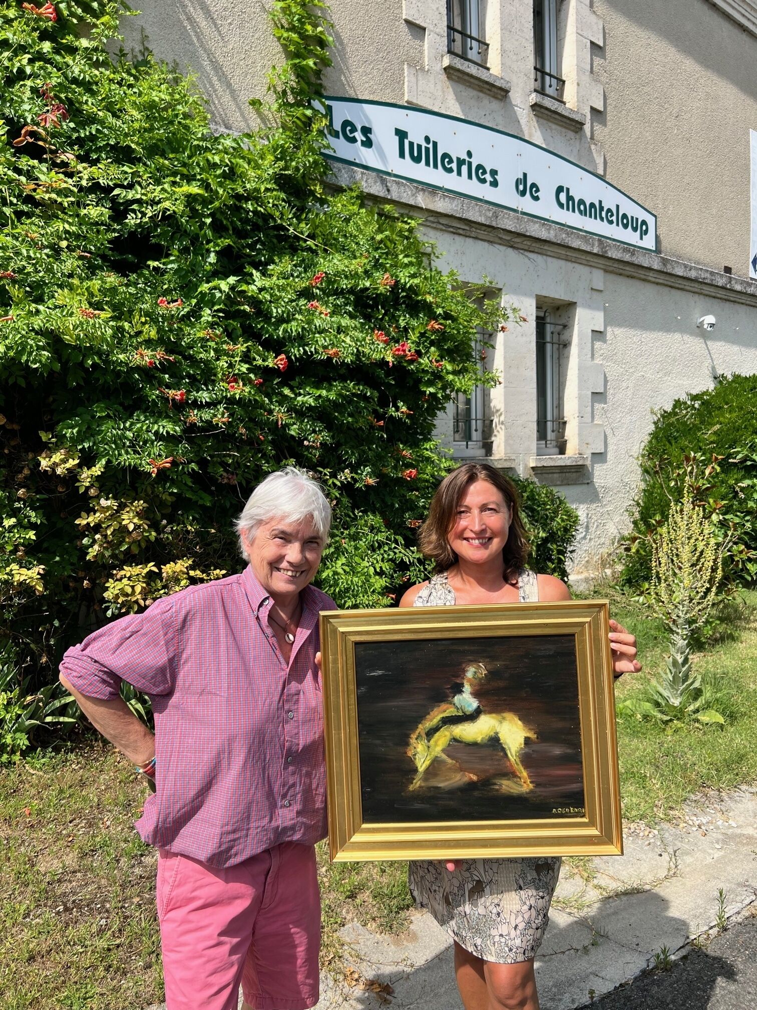 Two individuals holding a framed artwork in front of a building with a sign that reads 'Les Tuileries de Chanteloup.'