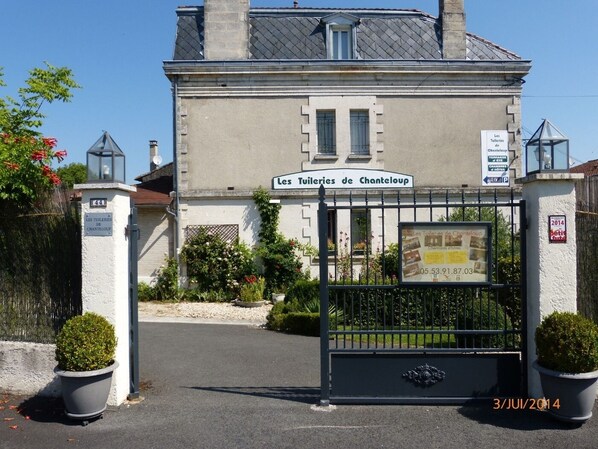 Una entrada con puerta metálica a un edificio identificado como 'Les Tuileries de Chanteloup', con un cartel que indica habitaciones de hotel y terrazas.
