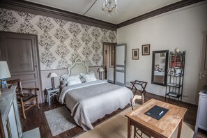 Un dormitorio con una cama grande, una mesita de noche de madera, una mesilla con lámpara, una silla de madera, una mesa de madera con una carpeta negra, un espejo y una estantería.