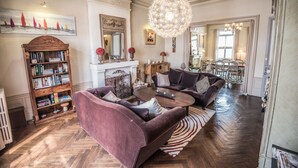 Een ruime woonkamer met een open haard, een grote kroonluchter en een boekenplank vol boeken.