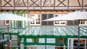 Un hotel con fachada verde y beige, amplias ventanas y zona de estar exterior con sombrillas.